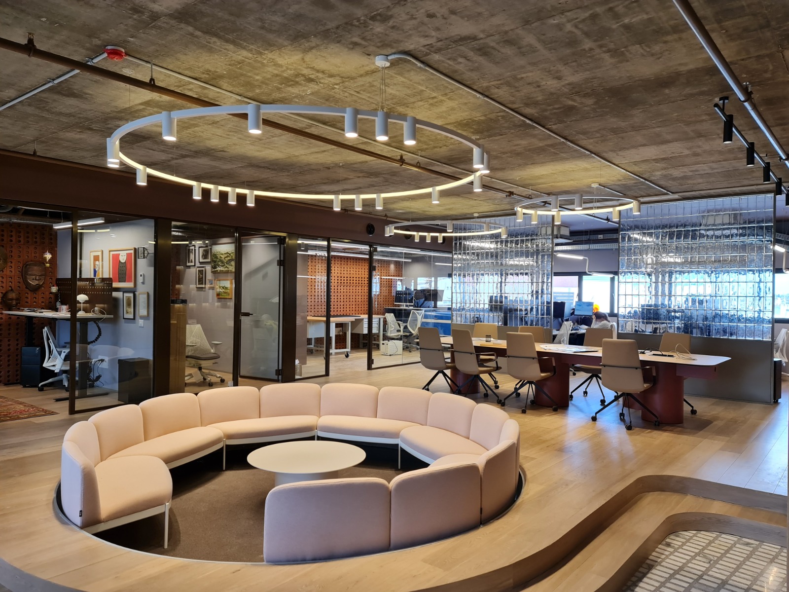 משרד מודרני עם תקרת בטון חשופה, שולחנות ישיבה ארוכים, חלל ישיבה עגול עם ספה רכה בגוון בז’, וגופי תאורה עגולים תלויים בעיצוב נקי ולבן היוצרים תאורה רכה ואחידה בחלל. ברקע מחיצות זכוכית בלוקים, קירות זכוכית וחללי עבודה פתוחים.