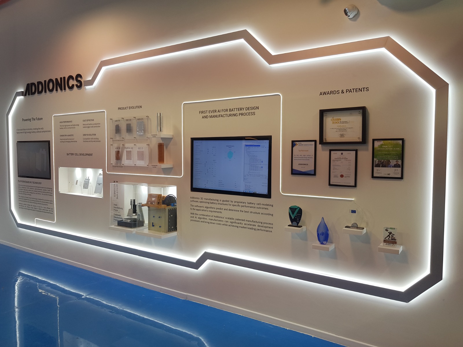 קיר תצוגה במשרדי חברת Addionics, הכולל פרסים, פטנטים ומידע על טכנולוגיית סוללות חדשנית. התאורה מדגישה את הצורה הגיאומטרית של הקיר באמצעות פסי לד שקועים, היוצרים קווים בהירים סביב האלמנטים ומעניקים מראה עתידני ונקי. הרצפה בצבע כחול מבריק מחזירה את האור ומחזקת את האפקט החזותי של התאורה.