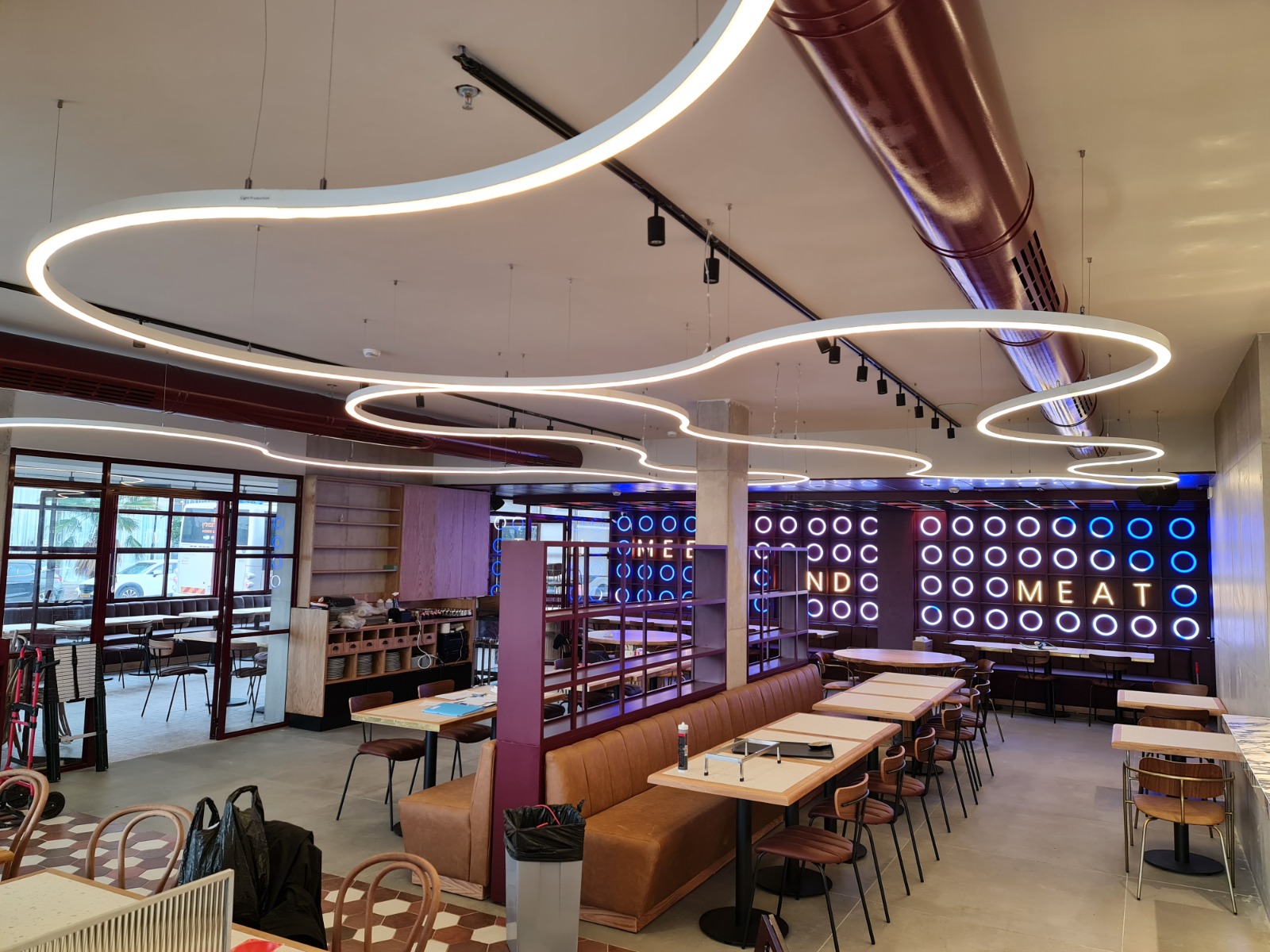 מסעדה בעיצוב מודרני עם פרופילי תאורה גמישים תלויים בתקרה בצורות אורגניות, המשרים תחושת זרימה ותנועה בחלל. ברקע קיר מואר בעשרות עיגולים בגווני כחול ולבן עם כיתוב מואר. הריהוט כולל ספות עור חומות, שולחנות עץ וכיסאות מתכת ועץ.